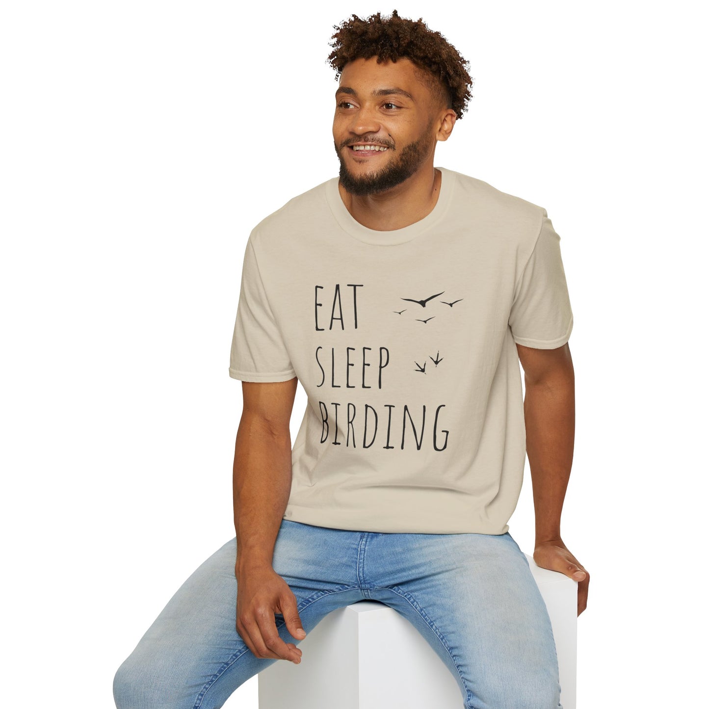 Man wearing a sand color t-shirt with 'Eat Sleep Birding' text, sitting on a white stool against a white background.
