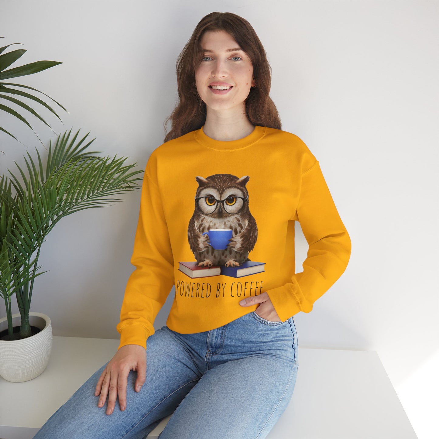 Woman wearing a yellow sweatshirt with an owl graphic and 'Powered by Coffee' text, sitting indoors.