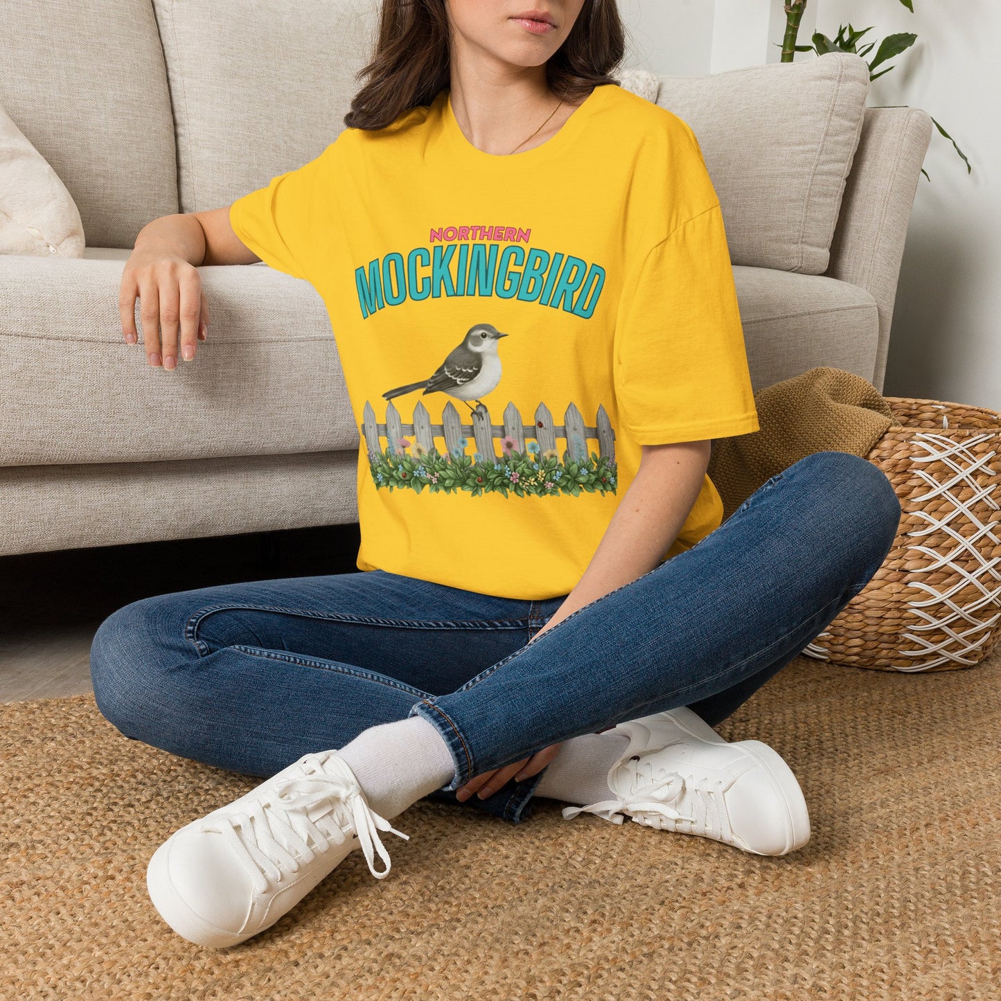 A woman wearing yellow t-shirt with a graphic print featuring the text 'NORTHERN MOCKINGBIRD' in teal and pink, an illustration of a mockingbird perched on a white picket fence with green foliage, with white background.