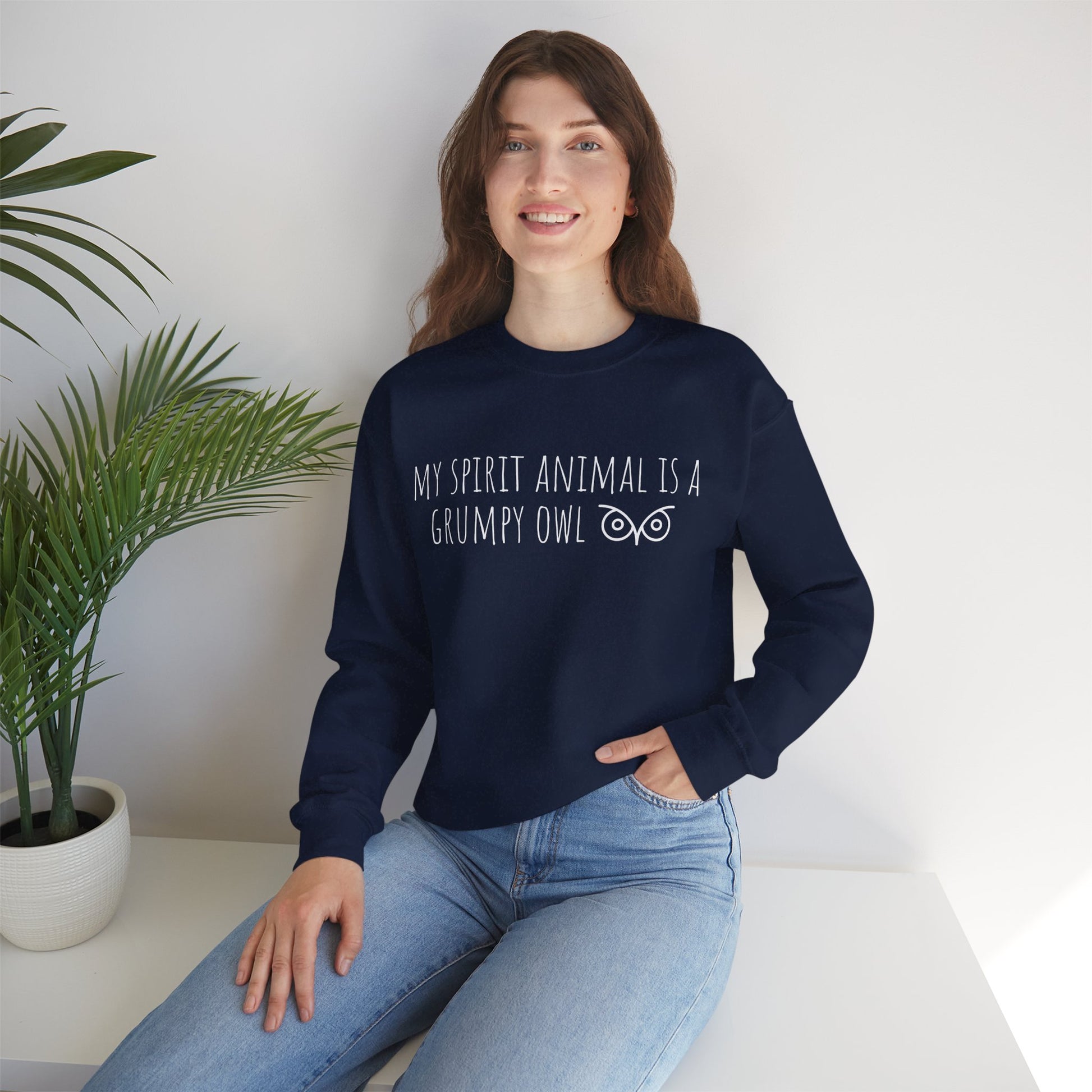 Woman wearing a navy blue sweatshirt with Owl text sitting on a white surface next to a plant.