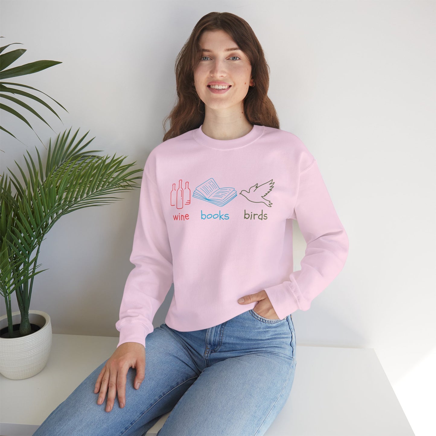 Person wearing a pink sweatshirt with 'wine books birds' design, sitting on a white surface with a plant in the background.