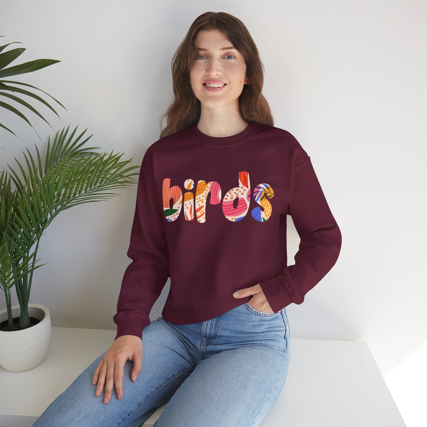 Person wearing a maroon sweatshirt with colorful 'birds' text, with a plant in the background.
