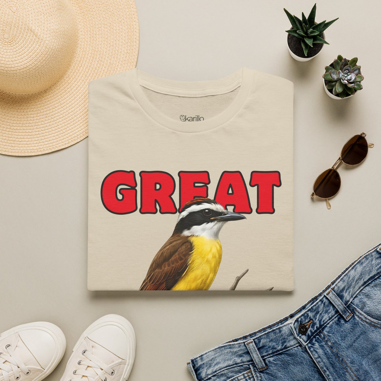Folded Beige t-shirt with a bird graphic and 'GREAT KISKADEE' text, surrounded by a hat, sunglasses, and shoes on a light surface.