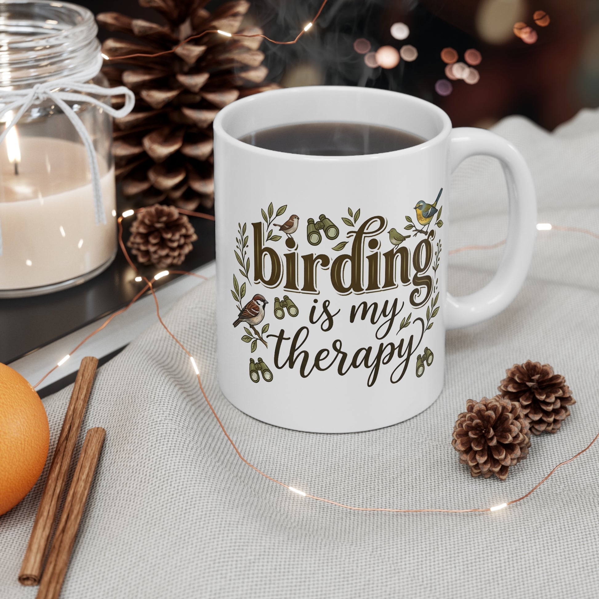 A white ceramic coffee mug with the phrase 'birding is my therapy' printed in brown script and decorated with small bird and leaf illustrations. The mug is filled with a hot beverage and styled on a light fabric surface with pinecones, fairy lights, a candle, cinnamon sticks, and an orange in the background.